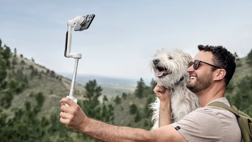 Ein Mann hält seinen Hund im Arm, während er ein Selfie mit dem integrierten Selfie-Stick von Insta360 Flow macht, mit Hügeln und Bäumen im Hintergrund.