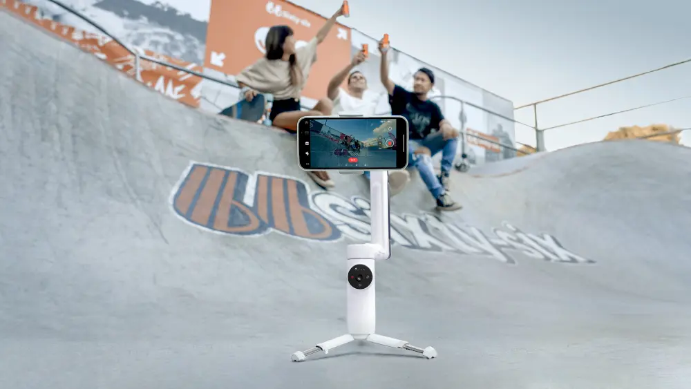Insta360 Flow mounted on the floor of a skatepark using it's built-in tripod. There are three skaters sat on the floor, being filmed by the smartphone and Flow.