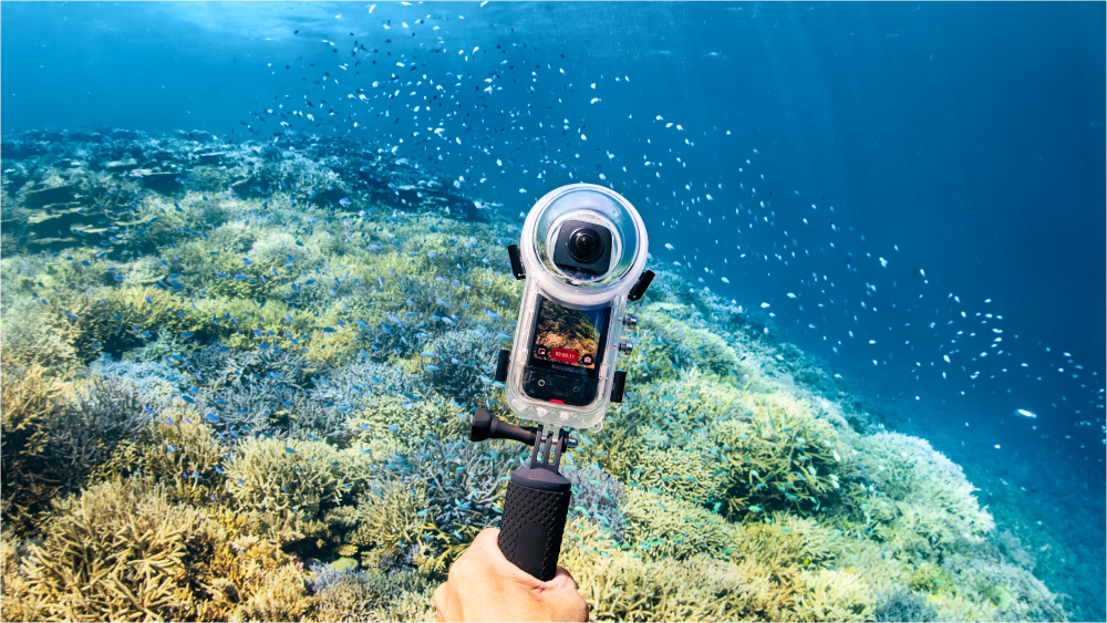 A diver's hand is visible holding Insta360 X3 in the Invisible Dive Case with coral and fish in the background.