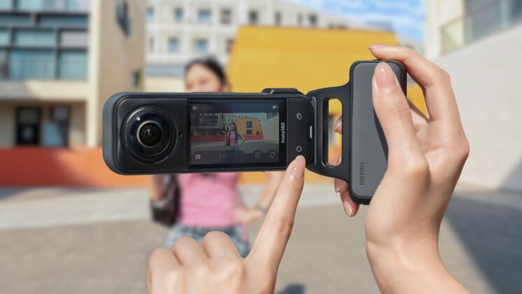 A pair of hands holding Insta360 X5 horizontally using the Insta360 Photography Grip. Their index finger on their left hand is about to press a button on the camera. They are taking a photo of a woman in a pink top in front of a bright yellow wall. 