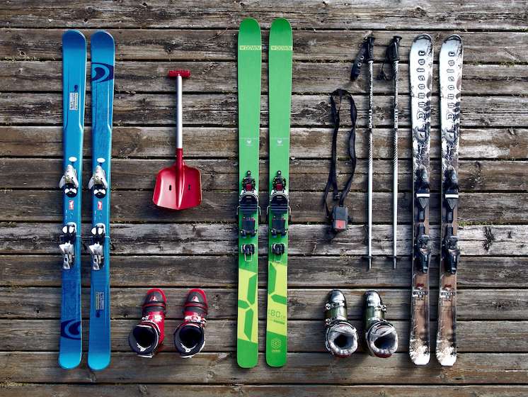 A picture of essential ski gear laid out on a deck. The ski equipment shown are three pairs of skis in blue, green and brown, a mini red shovel, two pairs of ski boots, a set of ski poles and a strap.