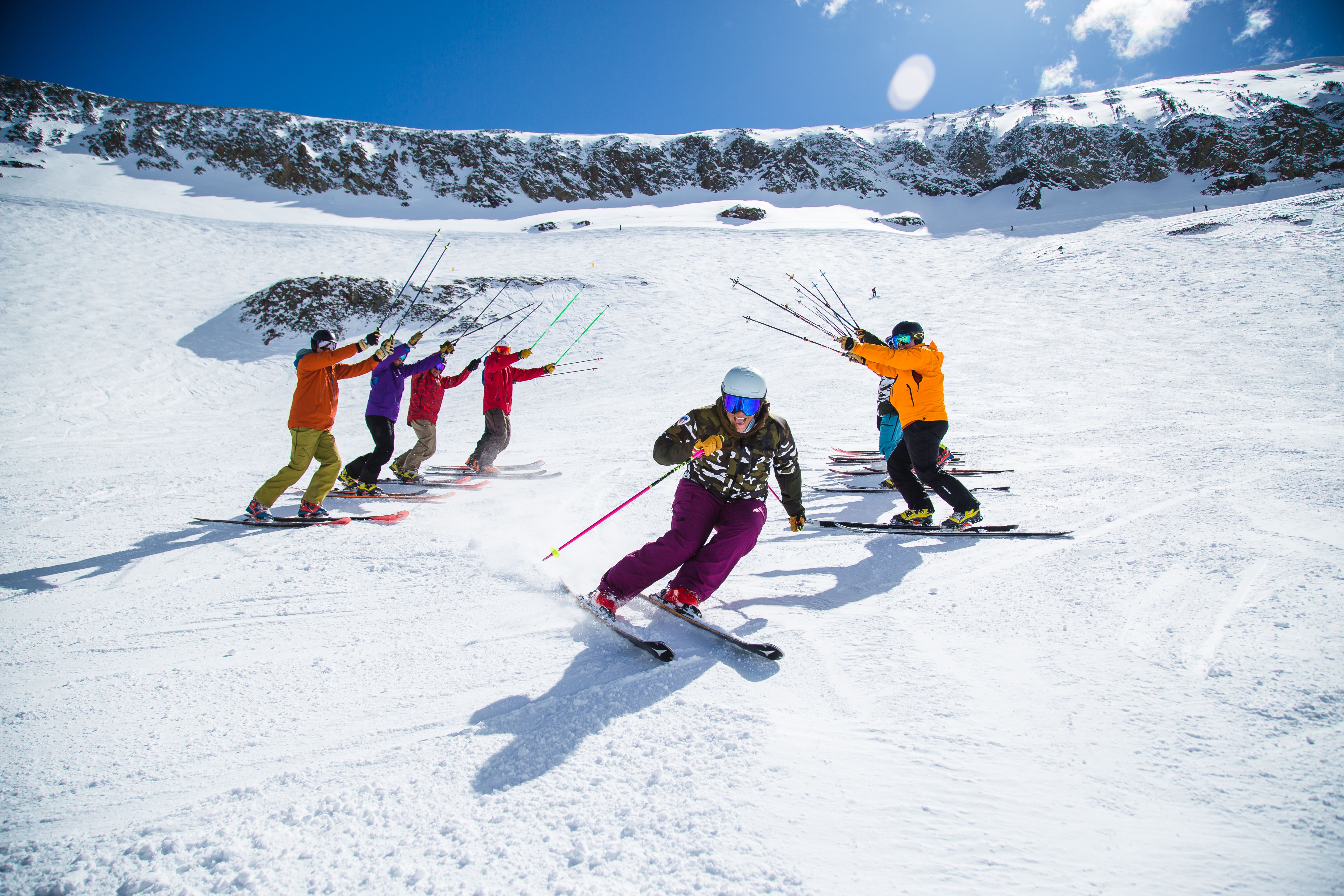 A skier going through a group of ski students. The students are in a line either side of the instructor and are holding up their ski poles to create an arch.