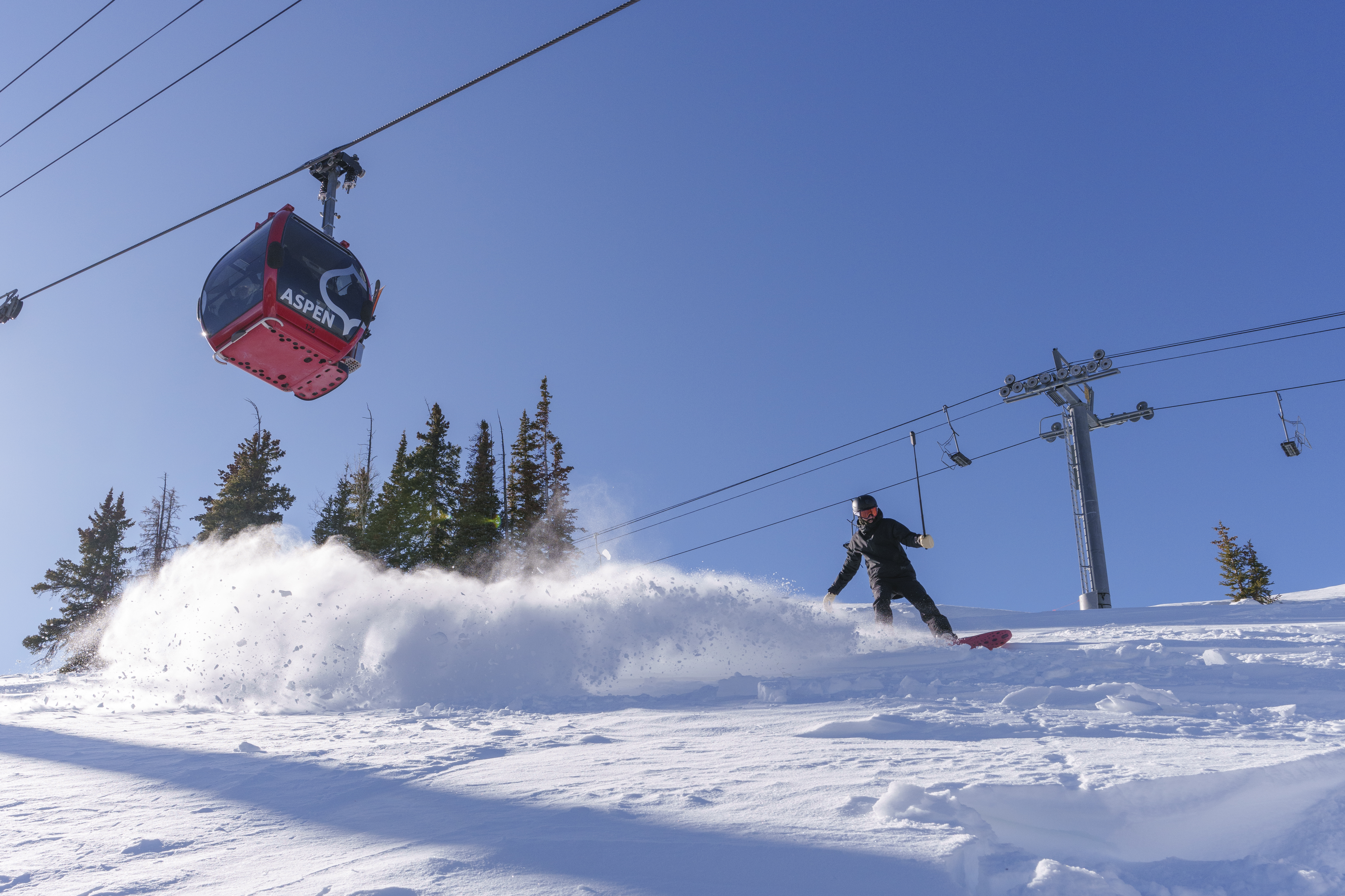 A snowboarder at Aspen Snowmass snowboarding in front of a ski lift. Insta360 X4 and the Insta360 Invisible Selfie Stick is out in front of themselves.