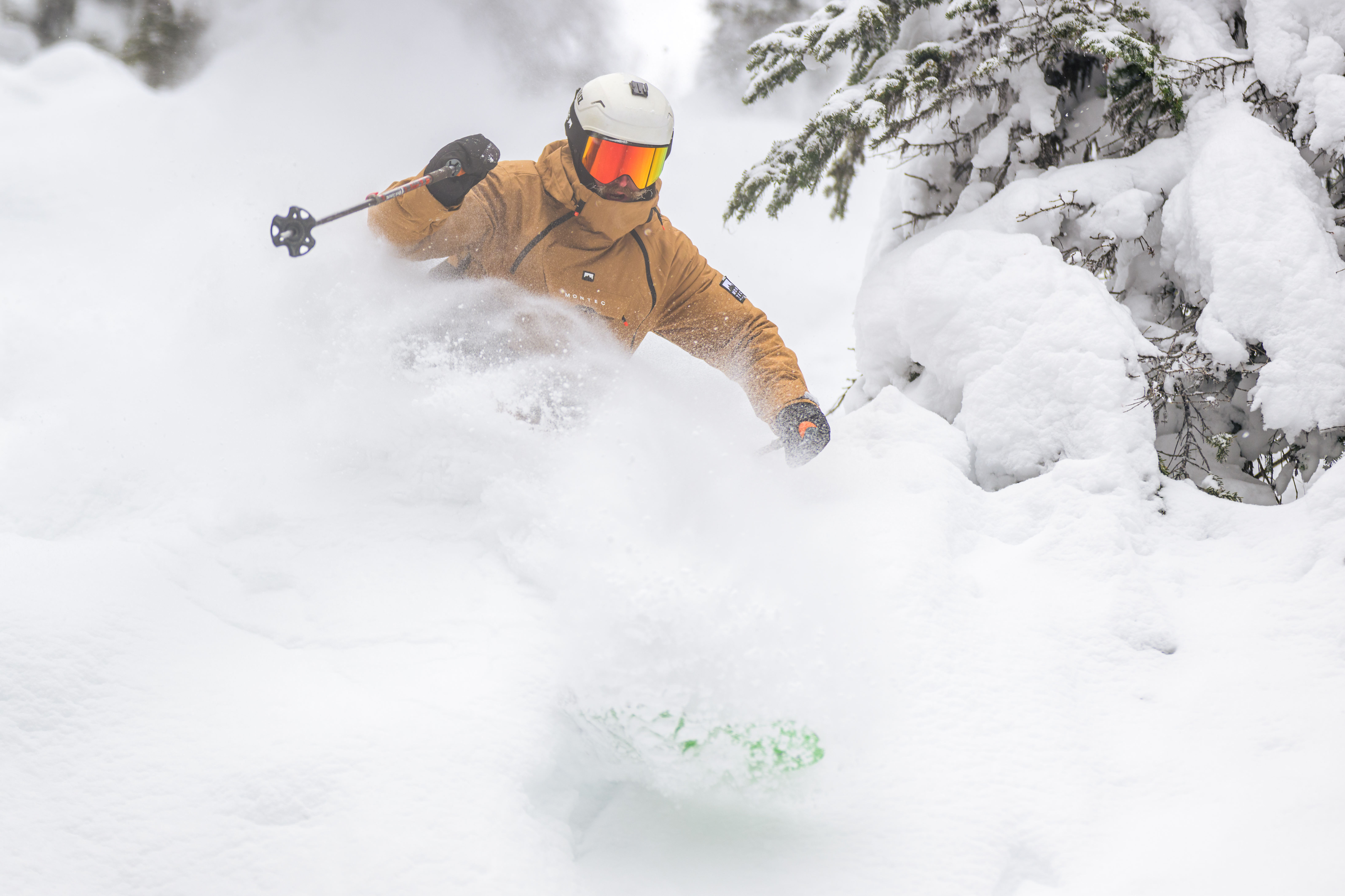 A skier in a light brown jacket and goggles skiing past a tree. Loose snow is covering his bottom half, indicating he is skiing fast.