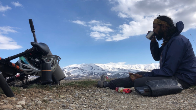 Kumar sitting on top of a mountain, sipping a drink, with his bike and bag on the floor