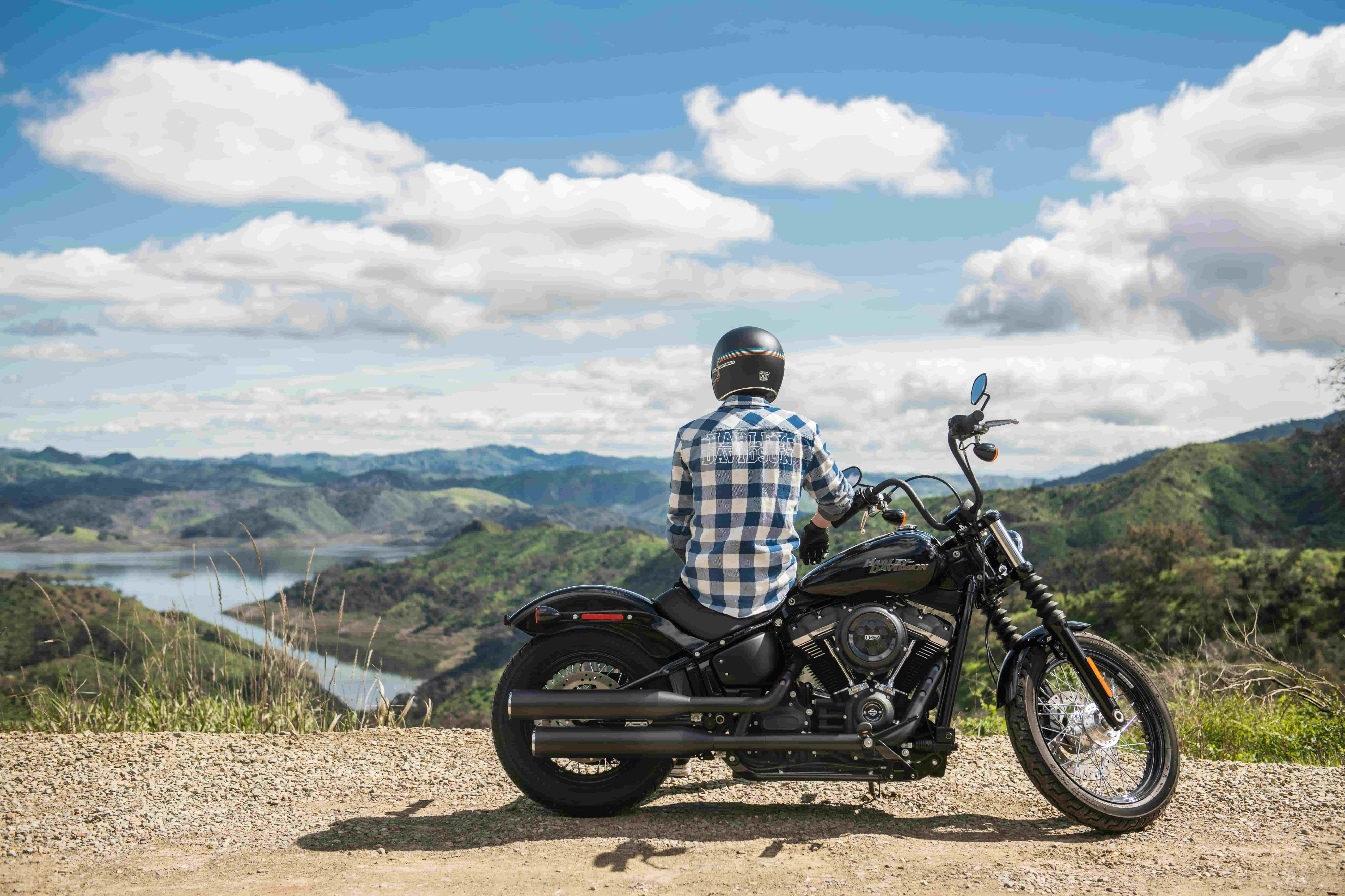 A motorcyclist parked on the side of the road, on his bike, looking at a view of mountains and a lake.
