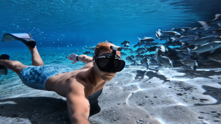 The tiny Insta360 GO 3S camera mounted on a man's goggles as he swims underwater with fish next to him