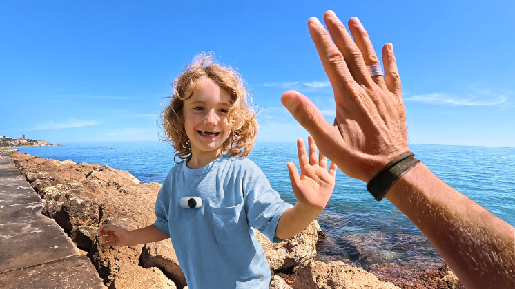 The tiny Insta360 GO 3S camera mounted on a child's chest using the Magnet Pendant, about to high five someone, with the sea in the background.