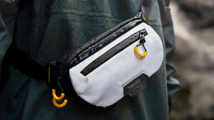 A man in a dark green coat wearing a white crossbody bag with orange zips on his back. His coat and the bag are wet with raindrops, demonstrating the bag is waterproof.