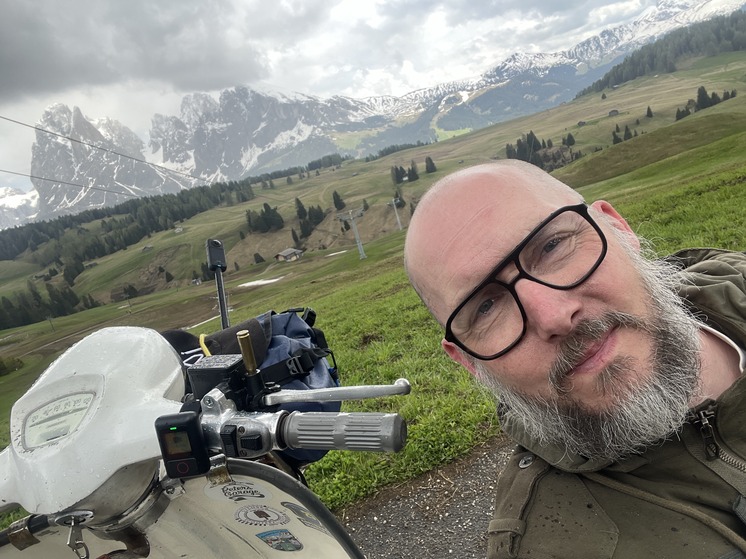 A man taking a selfie with his classic scooter in front of the Alps.