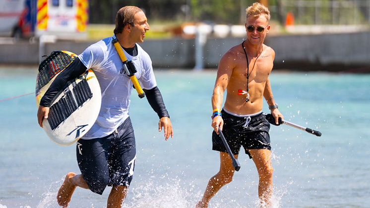Two surfers walking near the shoreline. One holding a surfboard and the other holding an Insta360 X5.